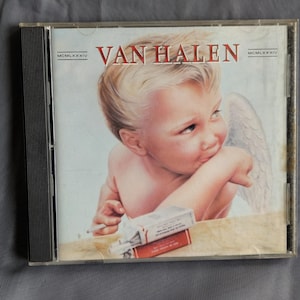 May include: A CD case featuring the band name "VAN HALEN" in red lettering. The album cover art depicts a cherubic figure with wings, holding a cigarette, and a pack of cigarettes. The case is in a clear plastic case.