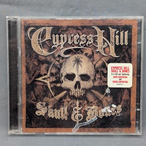 May include: A CD case featuring the Cypress Hill album "Skull & Bones." The cover art displays a skull with a marijuana leaf on its forehead, surrounded by bones and stars, with the band's name in a gothic font. A sticker indicates it's a 2-CD set.