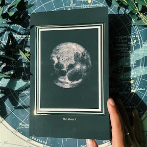 May include: A black framed print with a gold border featuring a detailed illustration of the moon on a dark green background. The text "The Moon" is printed below the image.