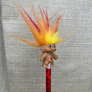 May include: A red and yellow troll pencil topper with a red gem on its chest. The troll has a spiky orange and red hair.