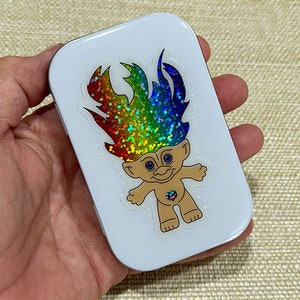 May include: A white metal tin with a rainbow glitter troll sticker. The troll has a heart-shaped gem on its chest.