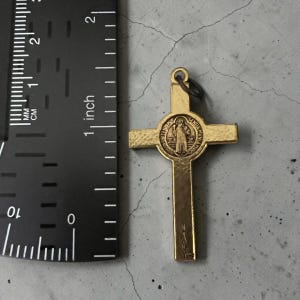 May include: A gold-toned metal crucifix pendant featuring a central circular medallion with a figure and text. The cross is positioned next to a black ruler, showing a total height of approximately 2.25 inches from top to bottom.