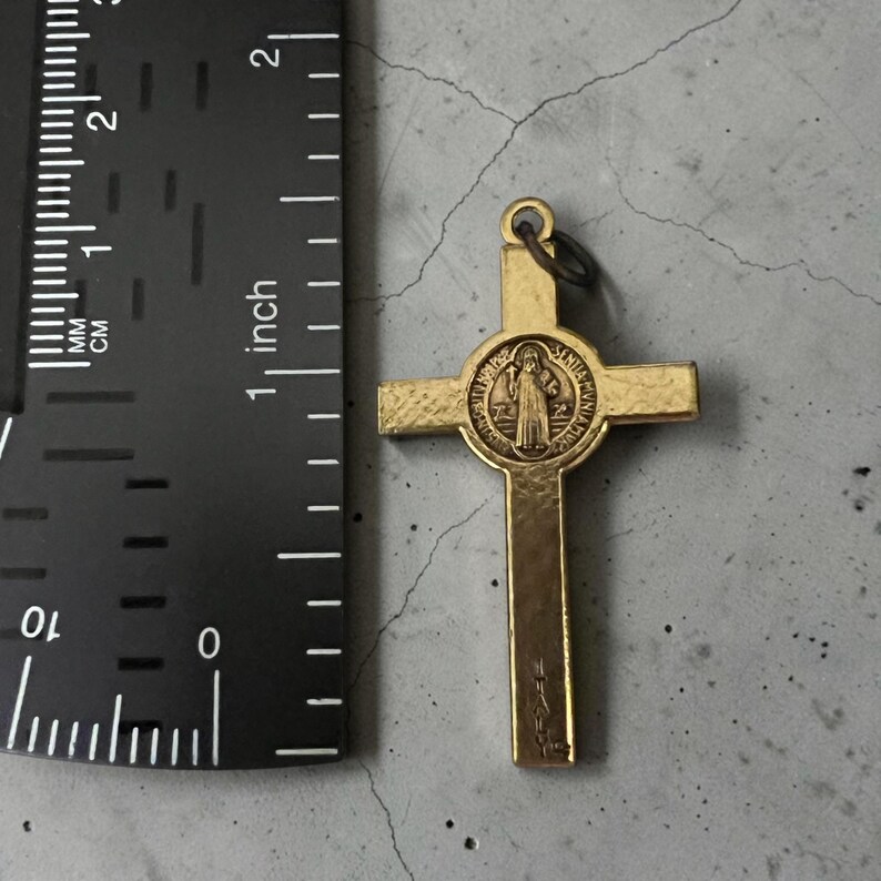 May include: A gold-toned metal crucifix pendant featuring a central circular medallion with a figure and text. The cross is positioned next to a black ruler, showing a total height of approximately 2.25 inches from top to bottom.
