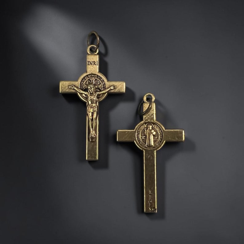 May include: Two gold-toned metal crucifix pendants featuring a central circular emblem. The left pendant depicts a figure on a cross with the letters INRI above. The right pendant shows a figure standing within the circular emblem.