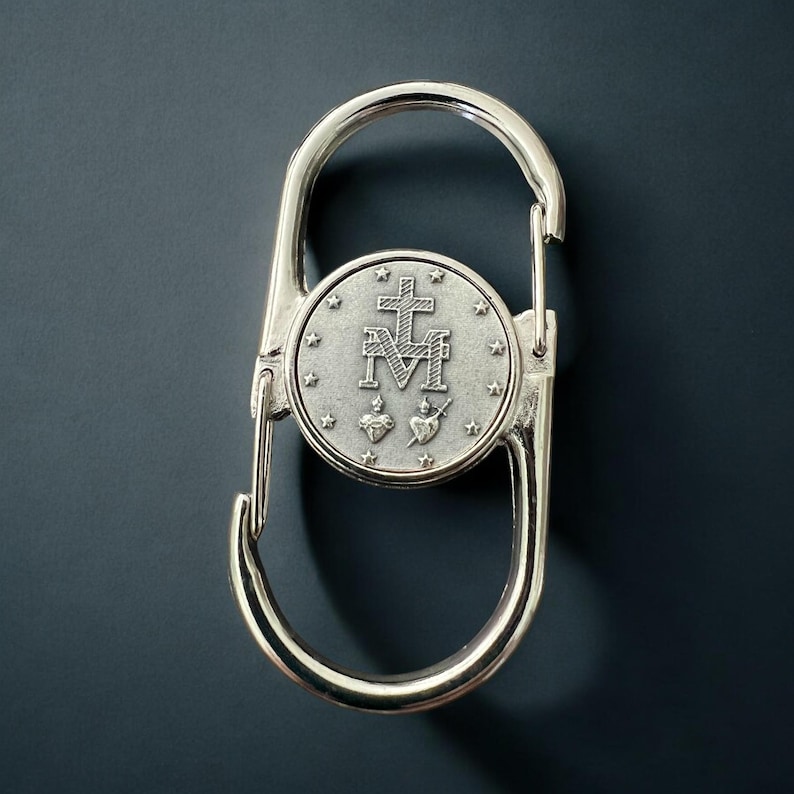 May include: Silver-toned metal keychain with a round charm featuring a religious symbol. The charm has a cross, the letters "M" and "M", and two hearts with a sword.
