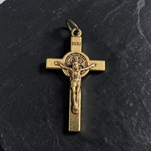 May include: A gold-colored crucifix pendant with the inscription "INRI" at the top. The cross features a detailed depiction of Jesus Christ. The pendant is designed with a loop for easy attachment to a chain or necklace.