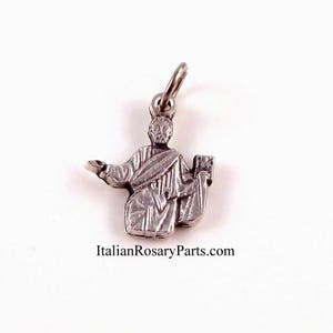 May include: Silver charm of a religious figure with a book in one hand and the other hand raised. The charm is for a rosary or necklace.