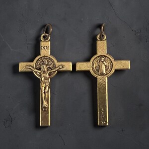 May include: Two gold-toned metal crucifix pendants. The left pendant features a figure of Jesus on the cross with the letters INRI above. The right pendant displays a circular Saint Benedict medal design and the word ITALY at the base.
