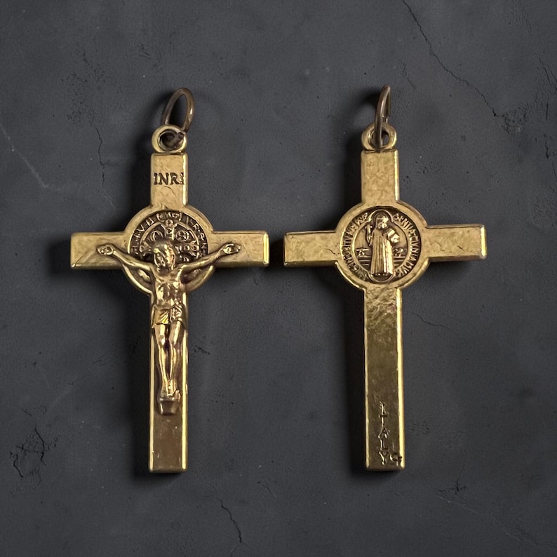 May include: Two gold-toned metal crucifix pendants. The left pendant features a figure of Jesus on the cross with the letters INRI above. The right pendant displays a circular Saint Benedict medal design and the word ITALY at the base.