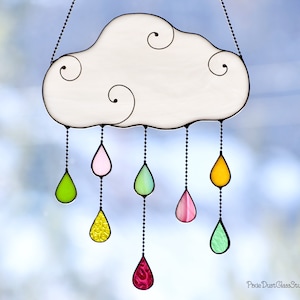 May include: A stained glass cloud with hanging teardrops. The cloud is white with black outlines and decorative swirls. The teardrops are various colors, including pink, green, and yellow, and are suspended by black beaded chains. The words "Pixie Dust Glass Studio" are in the bottom right corner.