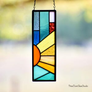 May include: A rectangular stained glass panel with a black border, featuring a sunburst design in shades of orange, yellow, blue, and green. The panel is suspended by a chain and includes the text "Pixie Dust Glass Studio".