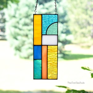 May include: A rectangular stained glass suncatcher featuring a geometric arrangement of yellow, blue, teal, orange, and white glass panels with varying textures, suspended by a black metal chain. The text Pixie DustGlassStudio is visible.