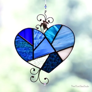 May include: Stained glass heart suncatcher in shades of blue, turquoise, and white. The heart is divided into geometric shapes and framed with black metal. A small turtle and decorative swirls adorn the top. The suncatcher hangs from a clear string.
