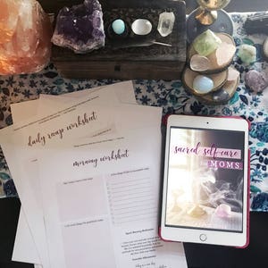 May include: A flat lay image of a white table with a pink Himalayan salt lamp, crystals, and a tablet displaying the text "sacred self-care for MOMS". There are also two printed worksheets on the table, one titled "daily recap worksheet" and the other titled "morning worksheet".
