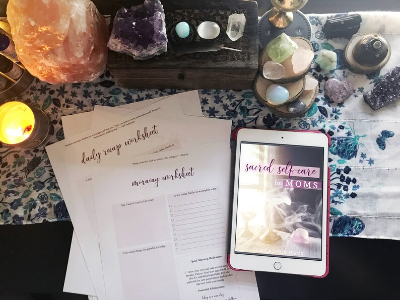 May include: A flat lay image of a white table with a pink Himalayan salt lamp, crystals, and a tablet displaying the text "sacred self-care for MOMS". There are also two printed worksheets on the table, one titled "daily recap worksheet" and the other titled "morning worksheet".