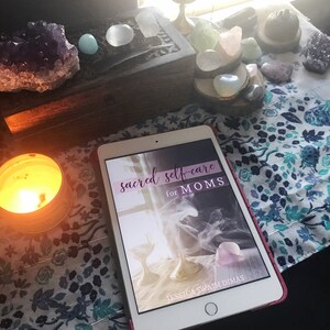 May include: A white tablet with a pink case displaying the cover of a book titled "sacred self-care for MOMS" by Jessica Swaim Dimas. The tablet is on a table with a lit candle, crystals, and a wooden box.