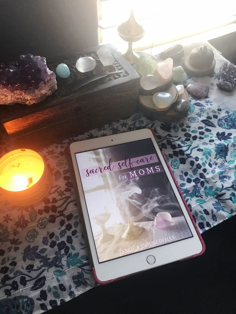 May include: A white tablet with a pink case displaying the cover of a book titled "sacred self-care for MOMS" by Jessica Swaim Dimas. The tablet is on a table with a lit candle, crystals, and a wooden box.