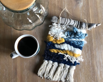 May include: A blue, white and yellow woven wall hanging with a driftwood branch and fringe.