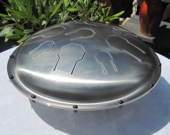 Könnte beinhalten: Ein silberfarbenes Handpan mit einem geometrischen Muster aus Vertiefungen. Das Handpan hat eine abgerundete Form und ist von einem Rand mit kleinen schwarzen Punkten umgeben.