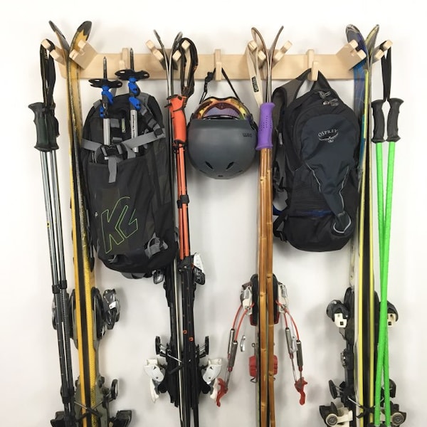Op de afbeelding: Een houten ski-rek met zes paar ski's, twee ski-stokken, twee rugzakken en een skihelm. Het rek is gemonteerd aan een witte muur.