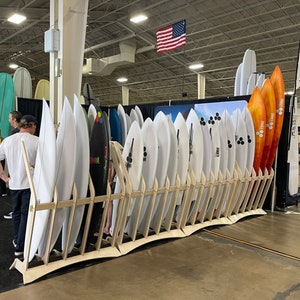 Puede incluir: Un estante de tablas de surf blancas y grises con una variedad de tablas de surf coloridas en el fondo. Las tablas de surf est&aacute;n dispuestas en fila y todas est&aacute;n apoyadas contra una pared.