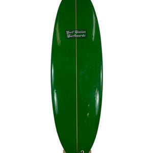 May include: A green surfboard with a white stripe down the center. The surfboard has a sticker on it that says "Surf Station Surfboards".