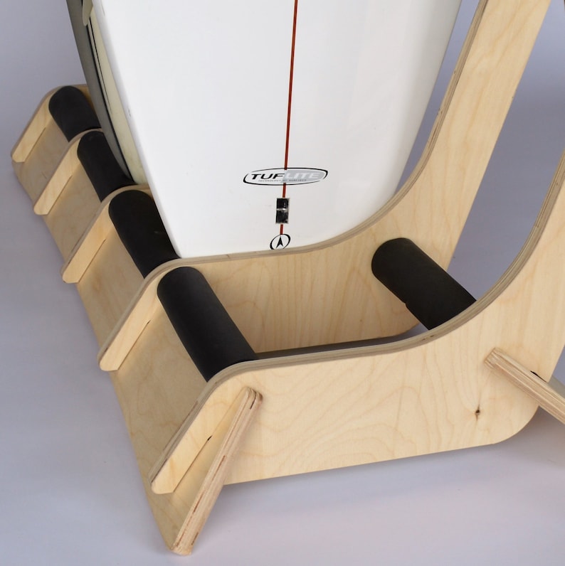 Puede incluir: Un soporte de tabla de surf de madera con acolchado de espuma negra. El soporte est&aacute; dise&ntilde;ado para sostener las tablas de surf en posici&oacute;n vertical y est&aacute; hecho de madera clara. El soporte tiene un dise&ntilde;o curvo y se muestra con una tabla de surf apoyada en &eacute;l.