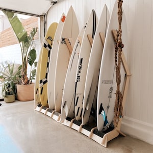 Puede incluir: Un estante de madera para tablas de surf con seis tablas de surf, incluyendo una tabla amarilla y negra con un patr&oacute;n a cuadros, una tabla blanca con texto negro que dice "Byer Surfboards" y una tabla blanca con texto negro que dice "The Grand Resort".