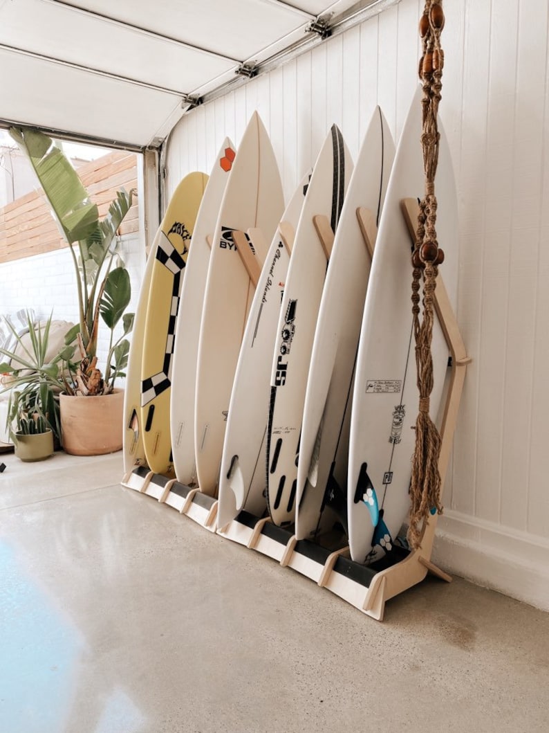 Puede incluir: Un estante de madera para tablas de surf con seis tablas de surf, incluyendo una tabla amarilla y negra con un patr&oacute;n a cuadros, una tabla blanca con texto negro que dice "Byer Surfboards" y una tabla blanca con texto negro que dice "The Grand Resort".