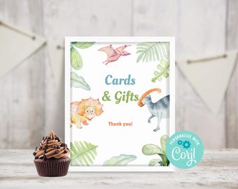 May include: A white frame with a sign that says "Cards & Gifts" with a dinosaur and a pterodactyl illustration. The sign is on a wooden surface with a chocolate cupcake.