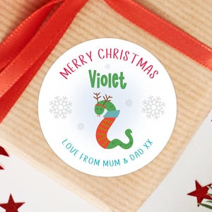 May include: A round white sticker featuring the text MERRY CHRISTMAS in red, the name Violet in green, and the text LOVE FROM MUM & DAD XX in teal. The design includes a green snake wearing brown reindeer antlers and a blue scarf, with snowflakes.