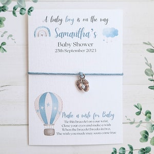 May include: A blue and white baby shower favour with a blue string bracelet and a charm of two tiny silver footprints. The card reads "A baby boy is on the way Samantha's Baby Shower 25th September 2023 Make a wish for Baby Tie this bracelet on your wrist, Close your eyes and make a wish When the bracelet breaks in two, The wish you made may soon come true"