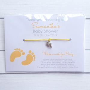 May include: A yellow string bracelet with a silver baby feet charm. The bracelet is attached to a white card with the text "Thank you for coming to Samantha's Baby Shower 27th October 2019" and "Make a wish for Baby... Tie this bracelet on your wrist, Close your eyes and make a wish When the bracelet breaks in two. The wish you made may soon come true."