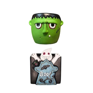 May include: Two Halloween-themed ceramic planters. The top planter is shaped like Frankenstein's monster with green skin, black hair, and stitched details. The bottom planter features a ghost with the word "Boo!" on a tombstone-shaped design.