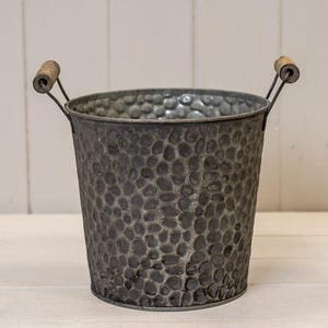 May include: A dark grey metal bucket with a textured, dimpled surface. It features two metal handles with wooden grips. This bucket is likely for decorative use or storage. The bucket's design has a rustic, vintage feel.