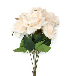 May include: A bunch of artificial cream-coloured roses. The roses have multiple layers of petals and are surrounded by green leaves. The stems are bundled and wrapped in brown material.