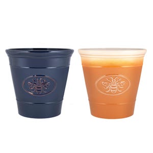 May include: Two ceramic plant pots. One is navy blue, the other is orange. Each pot features a bee emblem in the centre. The pots are cylindrical with a slightly flared top. The bee emblem is oval-shaped.