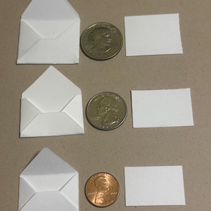 May include: Three miniature white envelopes, each next to a coin and a small white rectangle. Two quarters and a penny are shown. The envelopes are folded with a triangular flap. The rectangles are blank. The background is a neutral tan color.