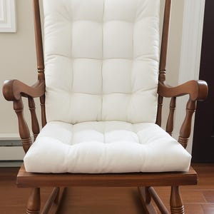 Puede incluir: Una mecedora de madera con un cojín blanco acolchado. La silla tiene un acabado marrón oscuro y un respaldo alto con tallas decorativas. El cojín es rectangular y se asegura con lazos blancos en la parte superior. La silla está sobre un piso de madera noble.