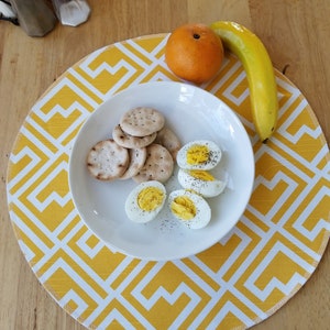 May include: A white plate with crackers and hard-boiled eggs on a yellow and white geometric placemat. There is a banana and an orange on the table.