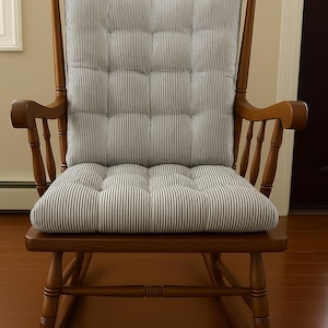 May include: A wooden rocking chair with a medium brown finish, featuring a tufted seat and back cushion. The cushion is upholstered in a white fabric with thin vertical grey stripes and is secured to the chair frame with small fabric ties.