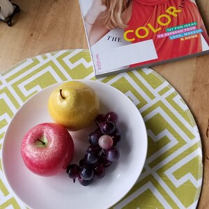 May include: A white bowl containing a yellow pear, a red apple, and a cluster of dark purple grapes. The bowl sits on a round placemat with a lime green and white geometric pattern. An InStyle magazine is visible with the text THE COLOR ISSUE.
