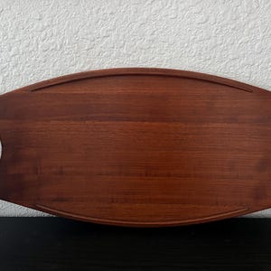 May include: A long, oval-shaped wooden serving tray with a rich, reddish-brown finish. The tray features a smooth, flat surface with a raised border and integrated cutout handles on each end, showcasing a natural wood grain pattern throughout.