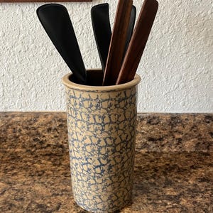 May include: A cylindrical ceramic utensil holder with a beige base and a blue sponge-painted mottled pattern. Inside the container are two black silicone spatulas and two brown wooden cooking utensils with flat, rectangular heads and long handles.