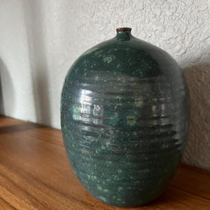 May include: A rounded ceramic vase with a narrow neck and a dark green glaze featuring mottled lighter green speckles and horizontal ribbed texture throughout the body. The surface has a glossy finish with subtle variations in color and tone.