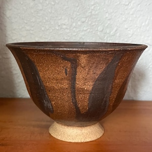May include: A ceramic bowl with a wide, flared rim and a tapered base. The exterior features a textured, speckled brown finish with dark brown vertical brushstroke patterns. The base is a lighter, unglazed beige color with a grainy texture.