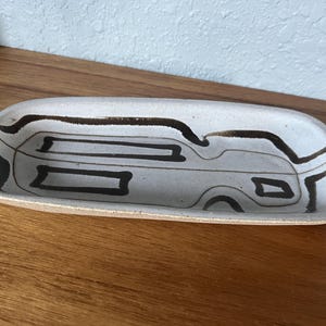 May include: A rectangular ceramic dish with a light gray base and a brown rim. The dish features a black abstract design. The dish is placed on a wooden surface.