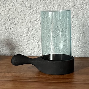 May include: A teal glass candle holder with a black wooden handle. The holder is sitting on a wooden surface.