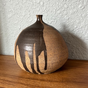 May include: A ceramic vase with a rounded body and a narrow neck. The vase is a speckled tan color with dark brown glaze dripping down the side. The vase sits on a wooden surface.