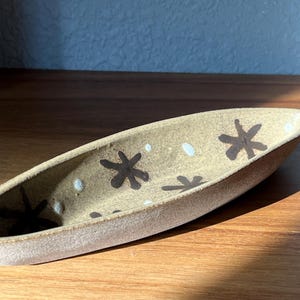 May include: A ceramic serving dish in the shape of a boat with a speckled brown glaze. The dish is decorated with white dots and black star-shaped designs.